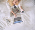 Eine Frau, welche mit Handy in der Hand auf dem Bett sitzt. Vor ihr ein PC. 